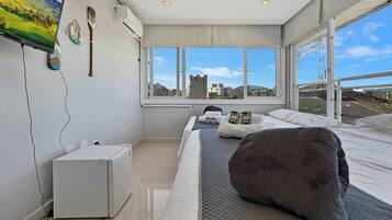 A modern bedroom with a large bed, a flat-screen TV, a small white cabinet, and a window with a city view.