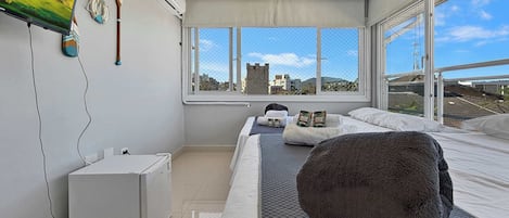 Quarto moderno com uma cama grande, televisão de tela plana, um armário branco pequeno e janela com vista para a cidade.