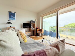 Una sala moderna con sofá, televisor, estantería y vista a un cuerpo de agua a través de amplios ventanales.
