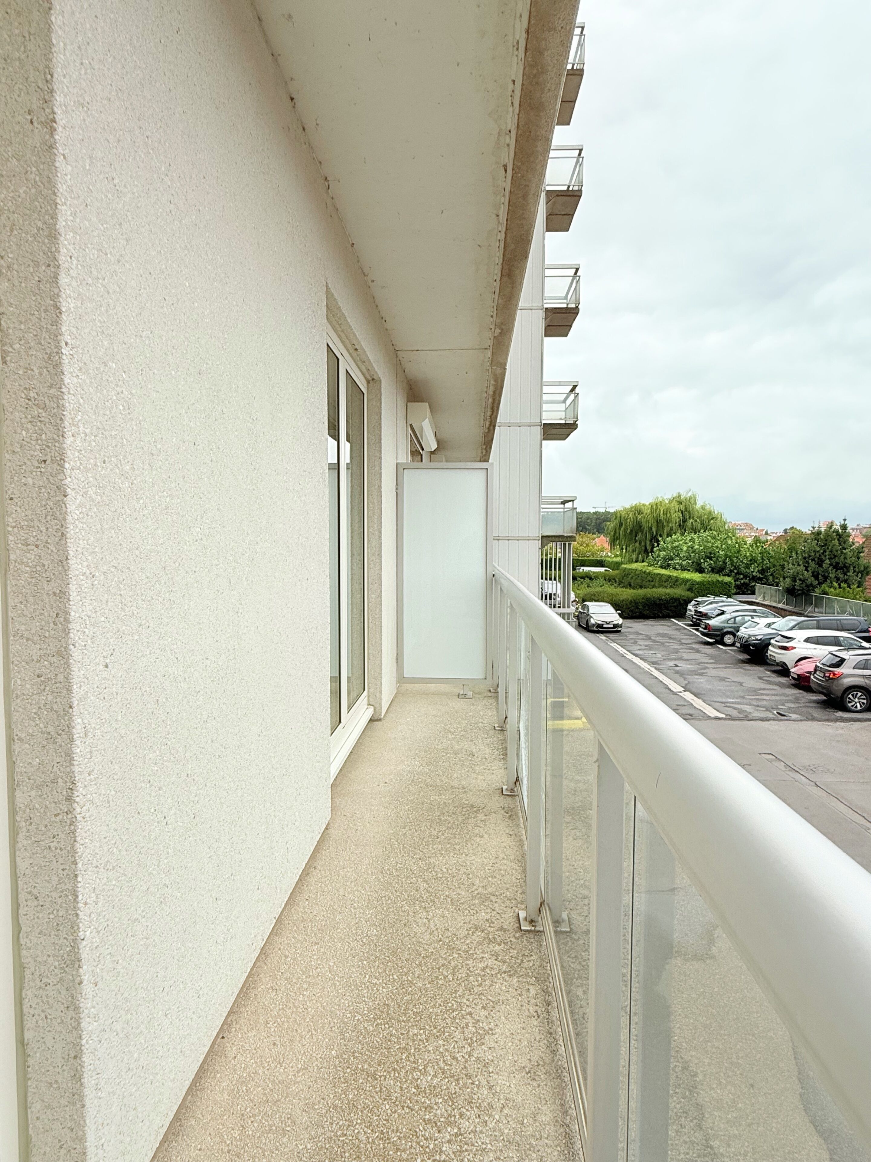Een smal balkon met een betonnen vloer, een witte balustrade en uitzicht op een parkeerplaats met auto's en groen op de achtergrond.