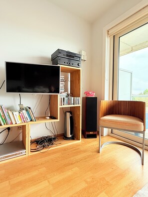 Ein modernes Wohnzimmer mit einem Flachbildfernseher, Holzregalen mit Büchern und elektronischen Geräten, einem Ledersessel und einem großen Fenster.