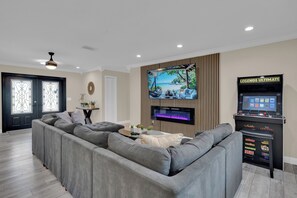 A living room with a large flat-screen TV mounted on a wooden panel, a gaming console, and a grey sectional sofa. - Waterfront Pool Home • Hot Tub • Private Golf Field (Tampa)