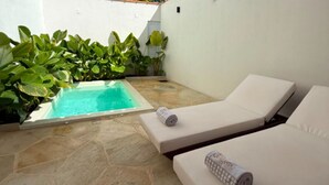 A small pool area with two white lounge chairs, one with a towel on it, and lush green plants around the pool.