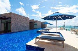 Ein schwebender Infinity-Pool auf dem Dach mit Liegestühlen und Sonnenschirmen, umgeben von moderner Architektur.