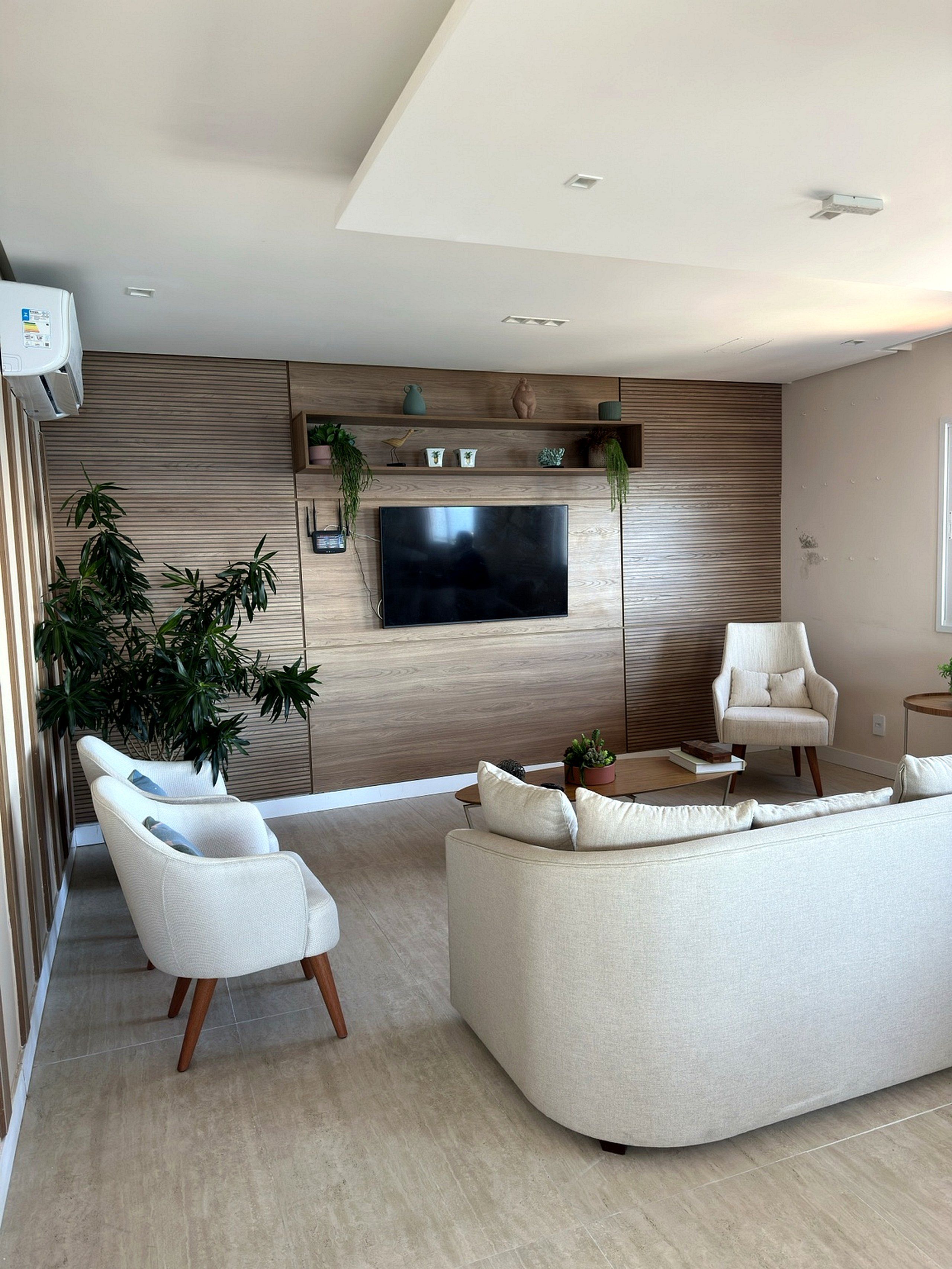 A modern living room with a flat-screen TV mounted on a wooden wall, a beige sofa, two white armchairs, and a coffee table with decorative items.