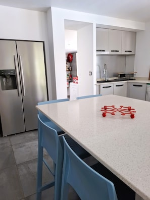 A modern kitchen with a stainless steel refrigerator, a white countertop, and blue chairs. - Villa Tartaruga - piscine privée, proche plage et centre ville (Sainte-Anne)