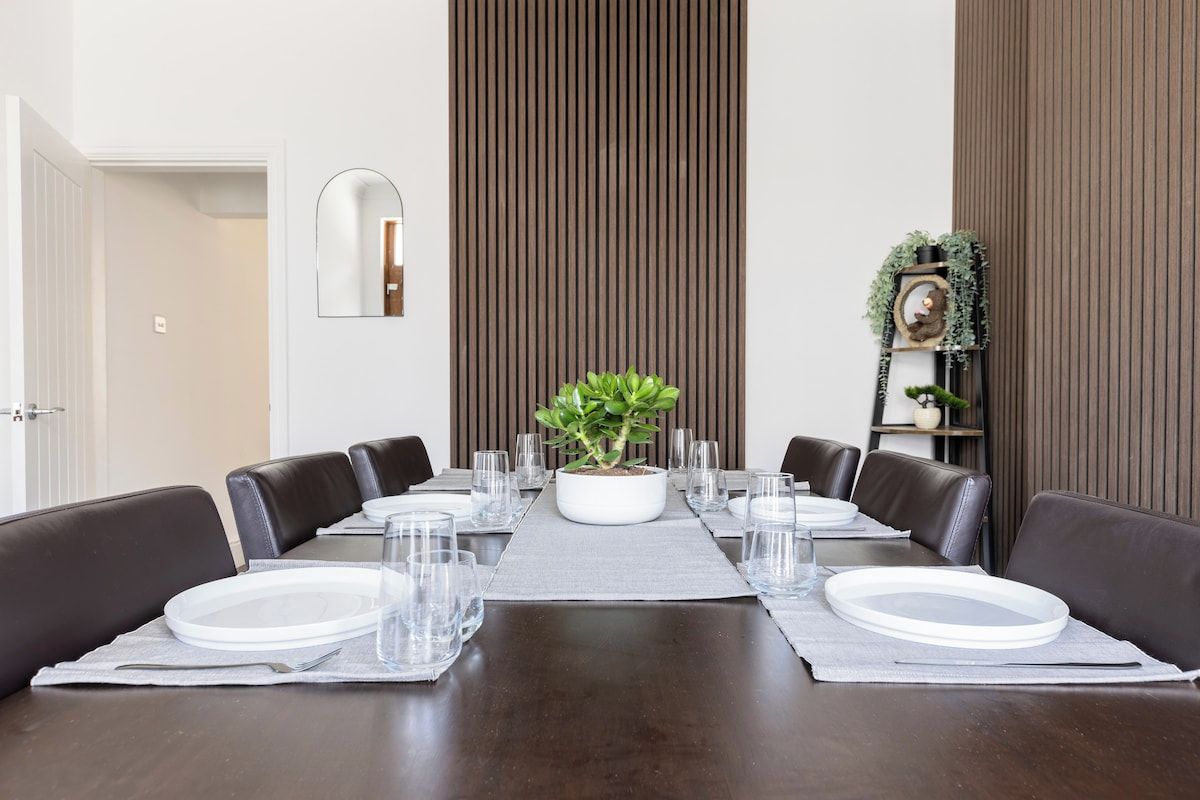 A dining room with a long table set for six, surrounded by dark leather chairs, a potted plant centerpiece, and a mirror on the wall.