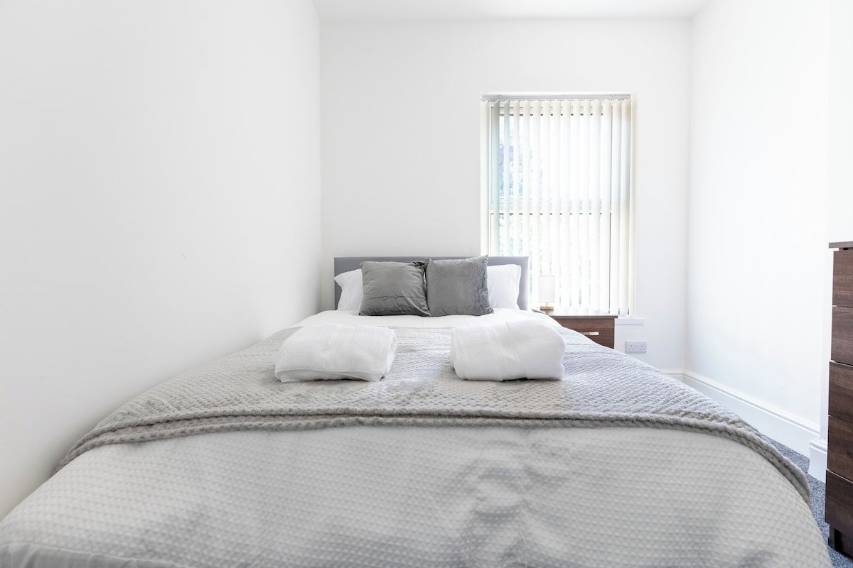 A bedroom with a bed, pillows, a bedside table, and a window with blinds.