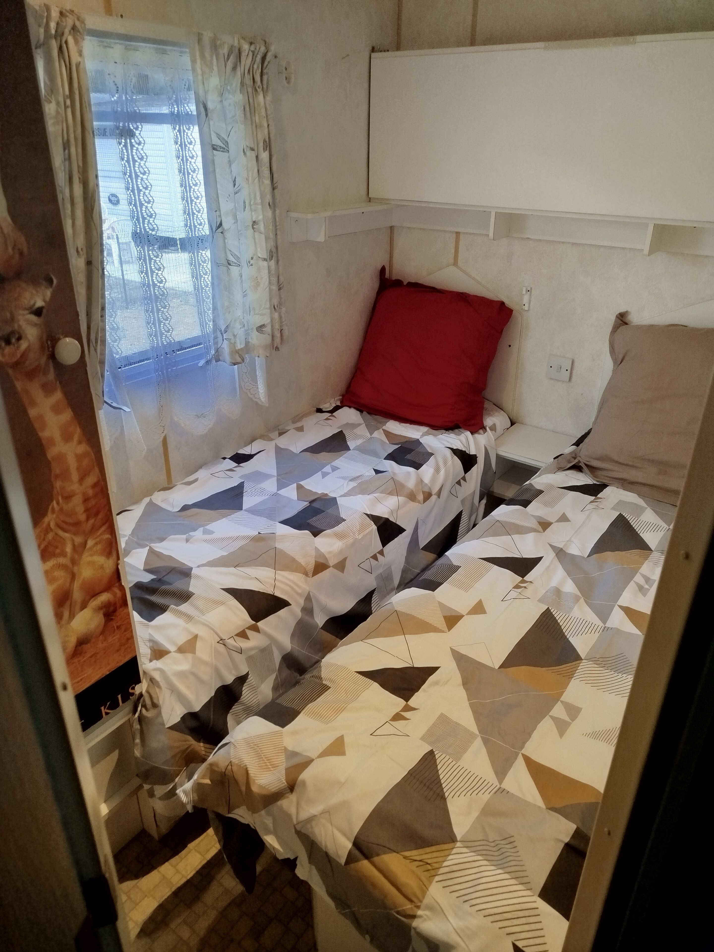 A small, single-bed room with a patterned bedspread, a red pillow, and a window with lace curtains.