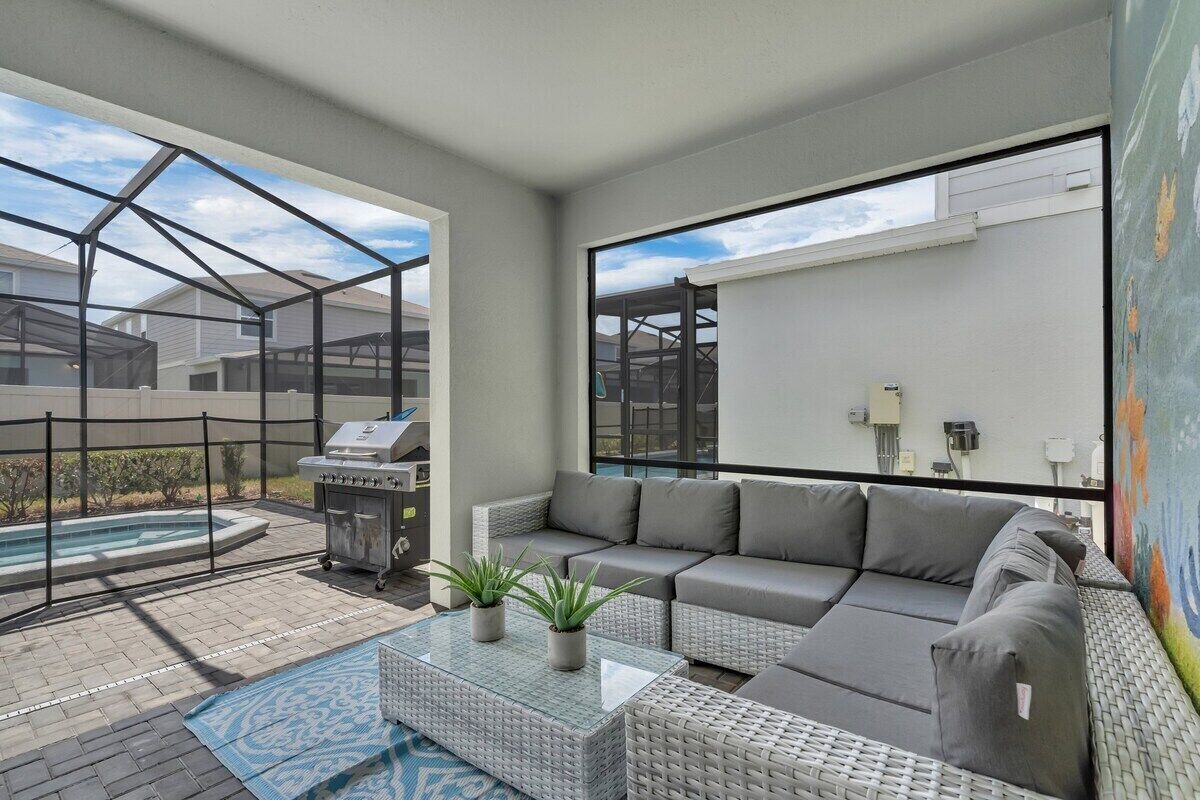 A modern outdoor living space with a glass coffee table, grey sofa, and a barbecue grill.