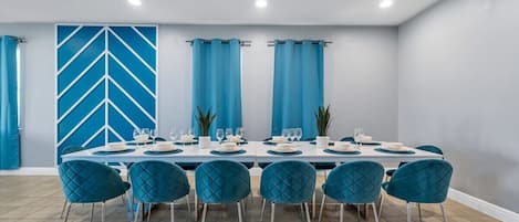 Un espace repas avec une longue table dressée pour six personnes, des chaises bleues et un motif géométrique sur le mur.