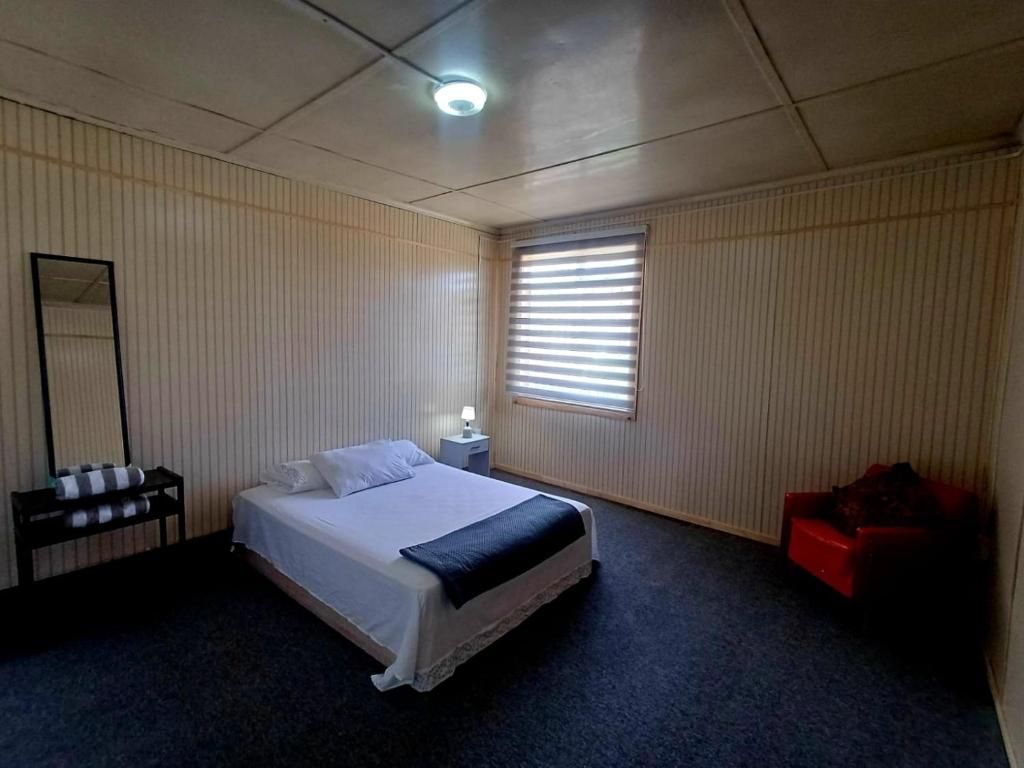Una camera d'albergo con un letto, un comodino, uno specchio, una finestra con tende e una sedia rossa.