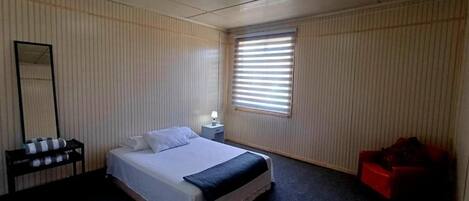 A hotel room with a bed, a nightstand, a mirror, a window with blinds, and a red chair.