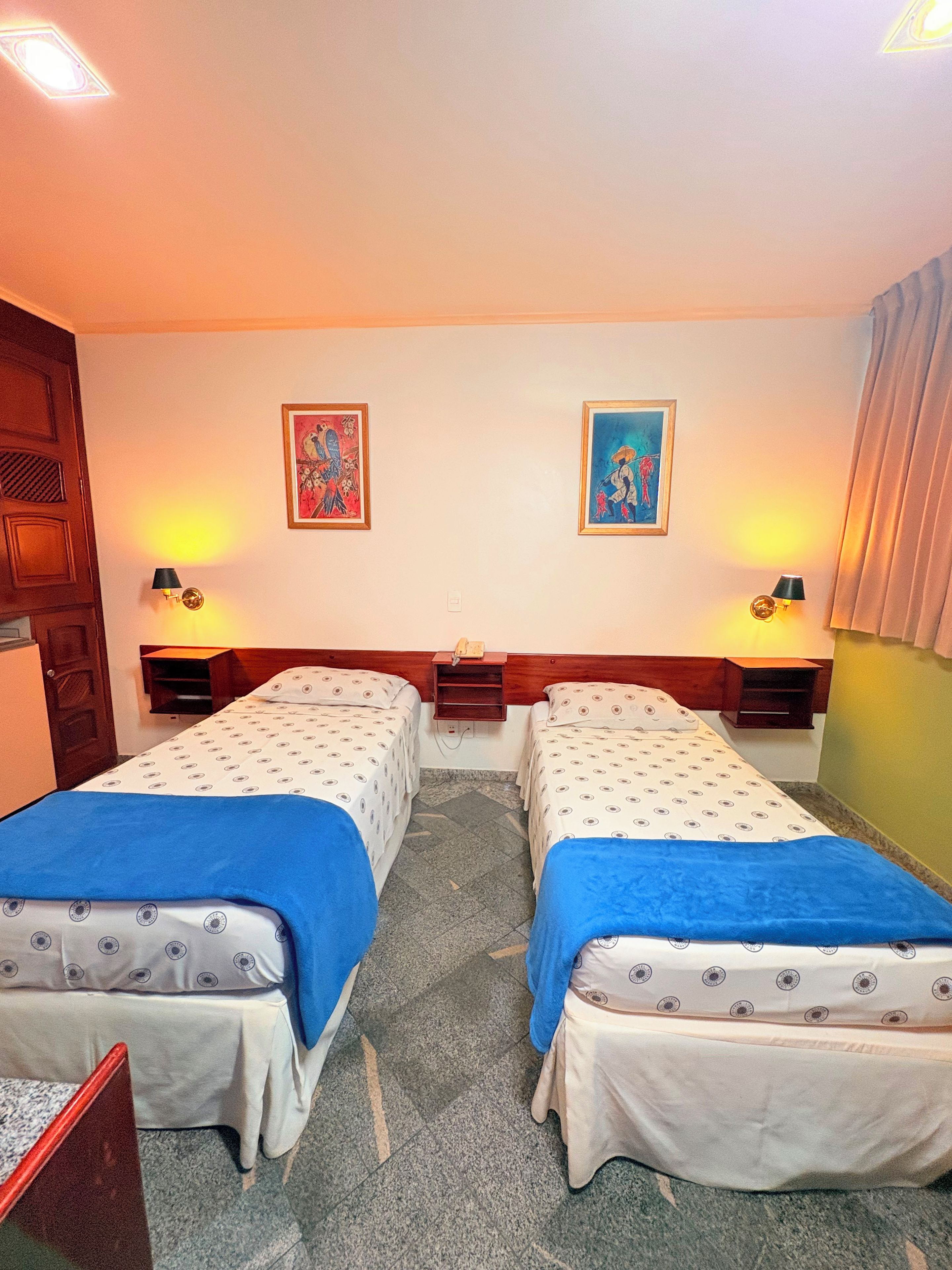 Habitación de hotel con dos camas, un cabecero de madera, una mesita pequeña, dos lámparas de pared y dos obras de arte enmarcadas.