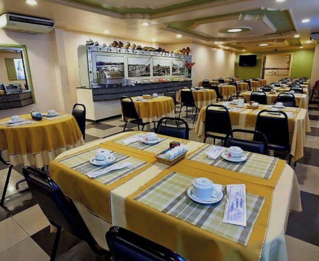 Uma área de refeições com mesas preparadas para o serviço, um balcão de buffet e um painel fixado na parede.