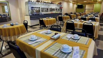 Uma área de refeições com mesas preparadas para o serviço, um balcão de buffet e um painel fixado na parede.