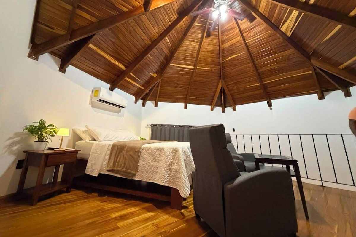 A bedroom with a wooden ceiling, a bed, a nightstand, a chair, and a small table with a lamp.