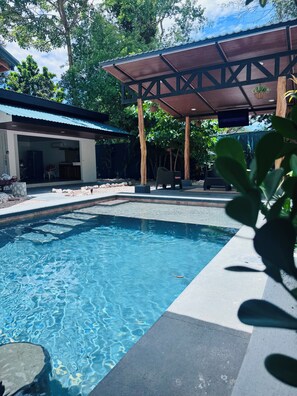 A swimming pool with a covered patio area, a building with a glass door, and surrounding greenery.