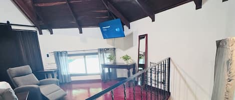 Amplia sala de estar con piso alfombrado en rojo, una escalera de madera y un televisor de pantalla plana montado en la pared.
