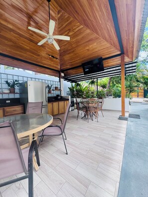 An outdoor patio with a wooden ceiling, a ceiling fan, a glass table, and chairs.