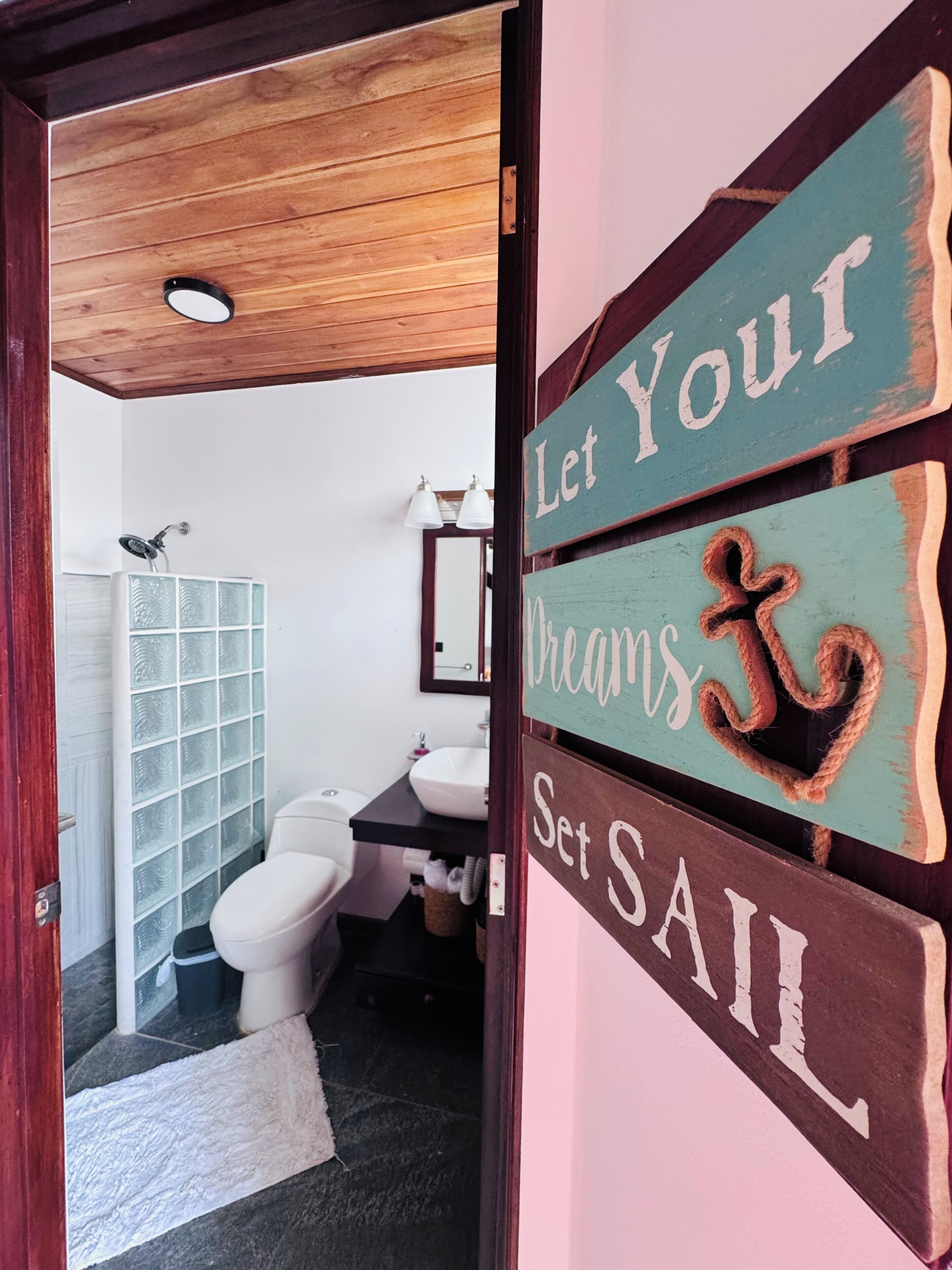 A bathroom with a shower, toilet, and sink. There is a wooden door with a sign that reads 'Let Your Dreams Set Sail.'