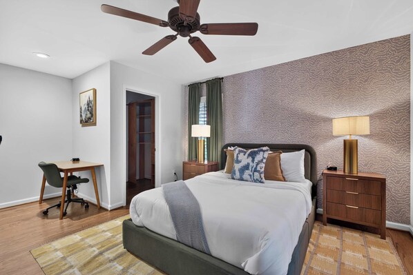 Habitación de hotel con cama, escritorio, silla, mesita de noche, lámpara y ventilador de techo.