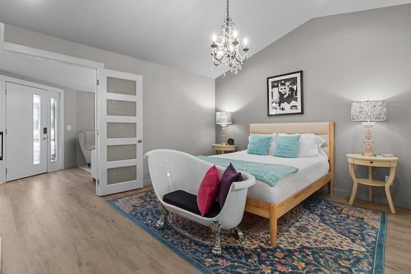 A bedroom with a bed, a white bathtub, a chandelier, and a door leading to another room.