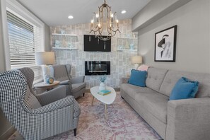 A living room with a television, a chandelier, a sofa, and patterned chairs. - The American Hotel (Williams Bay)