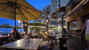 An outdoor dining area with tables and chairs, umbrellas, and a decorative lamp post. - Virginia Beach | Oceanfront Beach Retreat | Pool & Beach | 2BR (Virginia Beach)