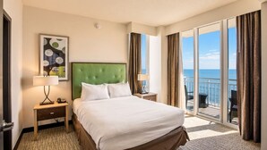A hotel room with a large bed, a nightstand, a lamp, a painting, and a balcony with a view of the sea. - Virginia Beach | Oceanfront Beach Retreat | Pool & Beach | 2BR (Virginia Beach)