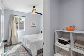 Ein Schlafzimmer mit einem Bett, einem Deckenventilator, einem Fenster mit Vorhängen, einem gerahmten Bild an der Wand und einem Regal mit gefalteter Kleidung und einer Kiste.