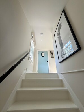 Eine Treppe führt zu einer Tür mit hellblauem Rahmen, ein Bild mit schwarzem Rahmen hängt an der Wand und ein Fenster mit Jalousien ist zu sehen.