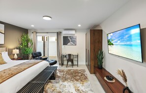 Una habitación de hotel moderna con cama, escritorio, silla y una gran pantalla de televisión mostrando una escena de playa.