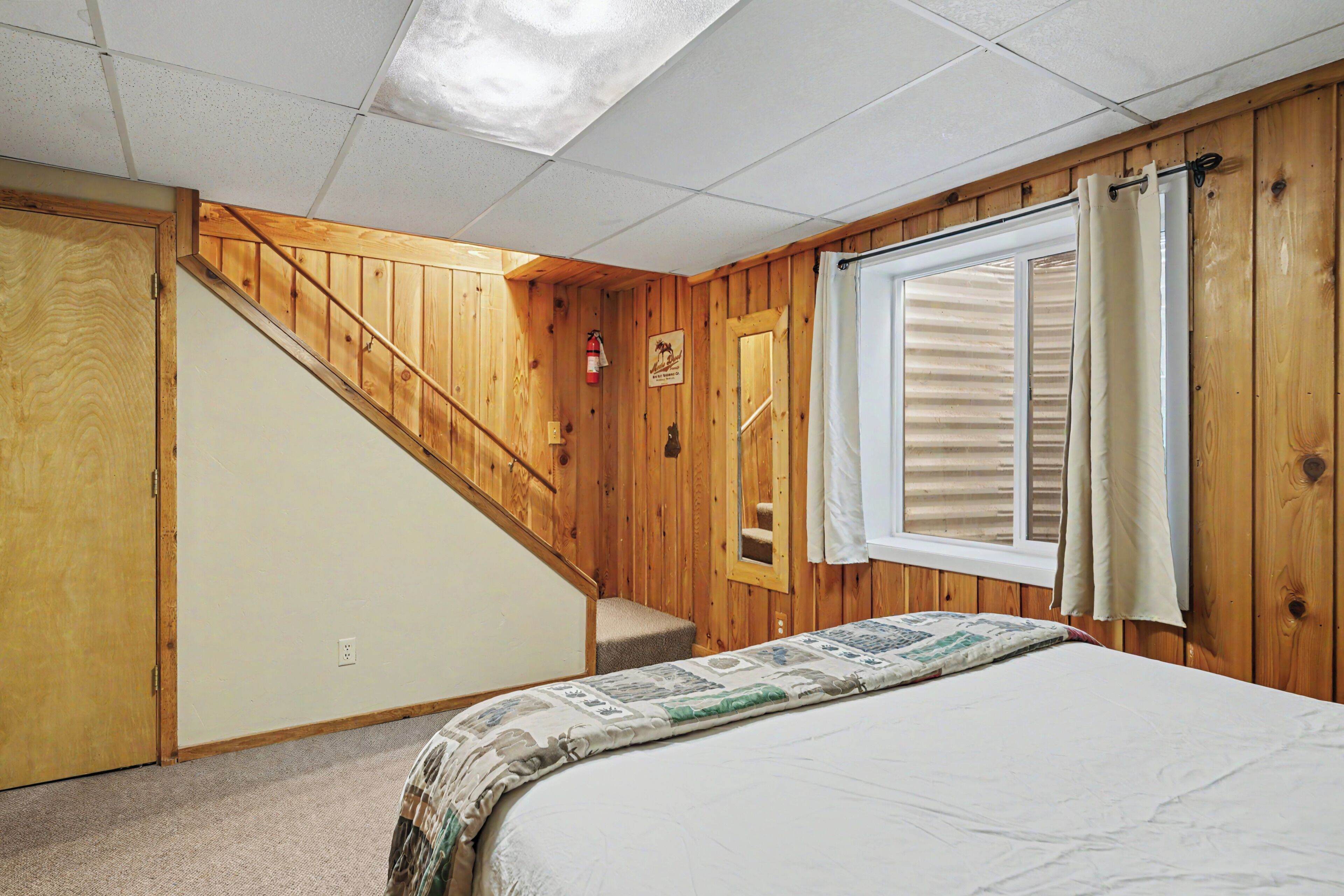 A bedroom with a wooden staircase, a bed with a patterned bedspread, a window with curtains, and a fire extinguisher on the wall.