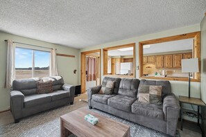 A living room with two grey sofas, a wooden coffee table, and a view of the kitchen through an open doorway. - Trapper 2 (West Yellowstone)