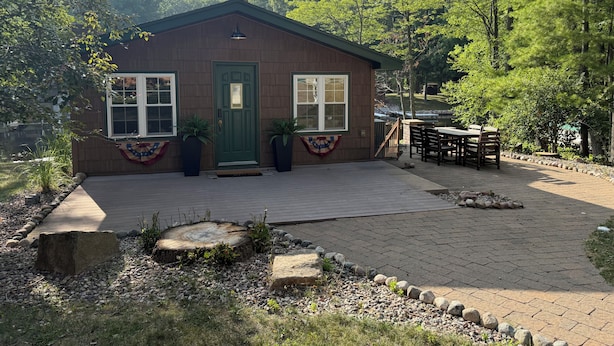 Eine kleine Holzhütte mit einer grünen Tür, zwei Fenstern und einer Terrasse mit Tisch und Stühlen.