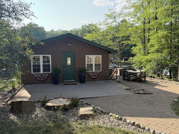 Une petite cabane en bois avec une porte verte, deux fenêtres et un espace patio comprenant une table et des chaises.