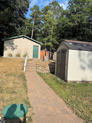 A pathway leading to a building with a green door, a shed, and a fenced area. - Cozy Lakefront Cottage — Private Dock & Fire Pit (Woodruff)