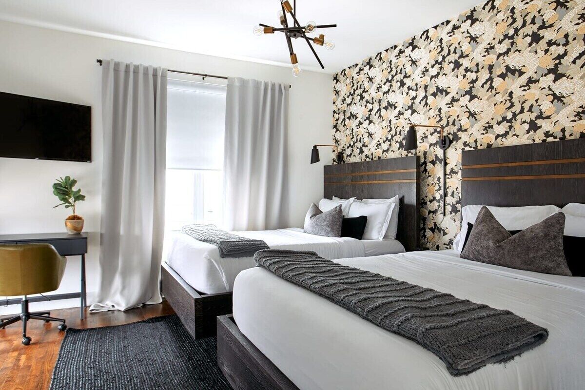 A hotel room with two beds, a desk, a chair, a potted plant, and a large window with curtains.