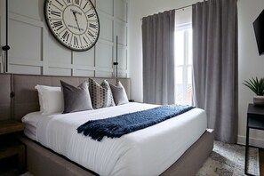 Ein Schlafzimmer mit einer großen Wanduhr, einem Bett mit blauer Tagesdecke und einem Fenster mit Vorhängen.