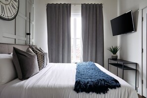 A bedroom with a bed, curtains, a wall-mounted TV, and a clock. - City Views In Hillsboro Village for 24 Guests, Garage, Walk to Vanderbilt And Boutique Shopping (Nashville)