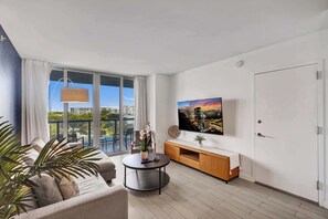 Uma sala de estar moderna com um sofá, uma mesa de centro redonda, uma TV na parede e uma varanda com vista para a cidade.