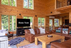 Ein gemütliches Cabin-Interieur mit einem großen Fernseher, Ledersofas, einem Holztisch und Blick auf den Wald durch die Fenster.