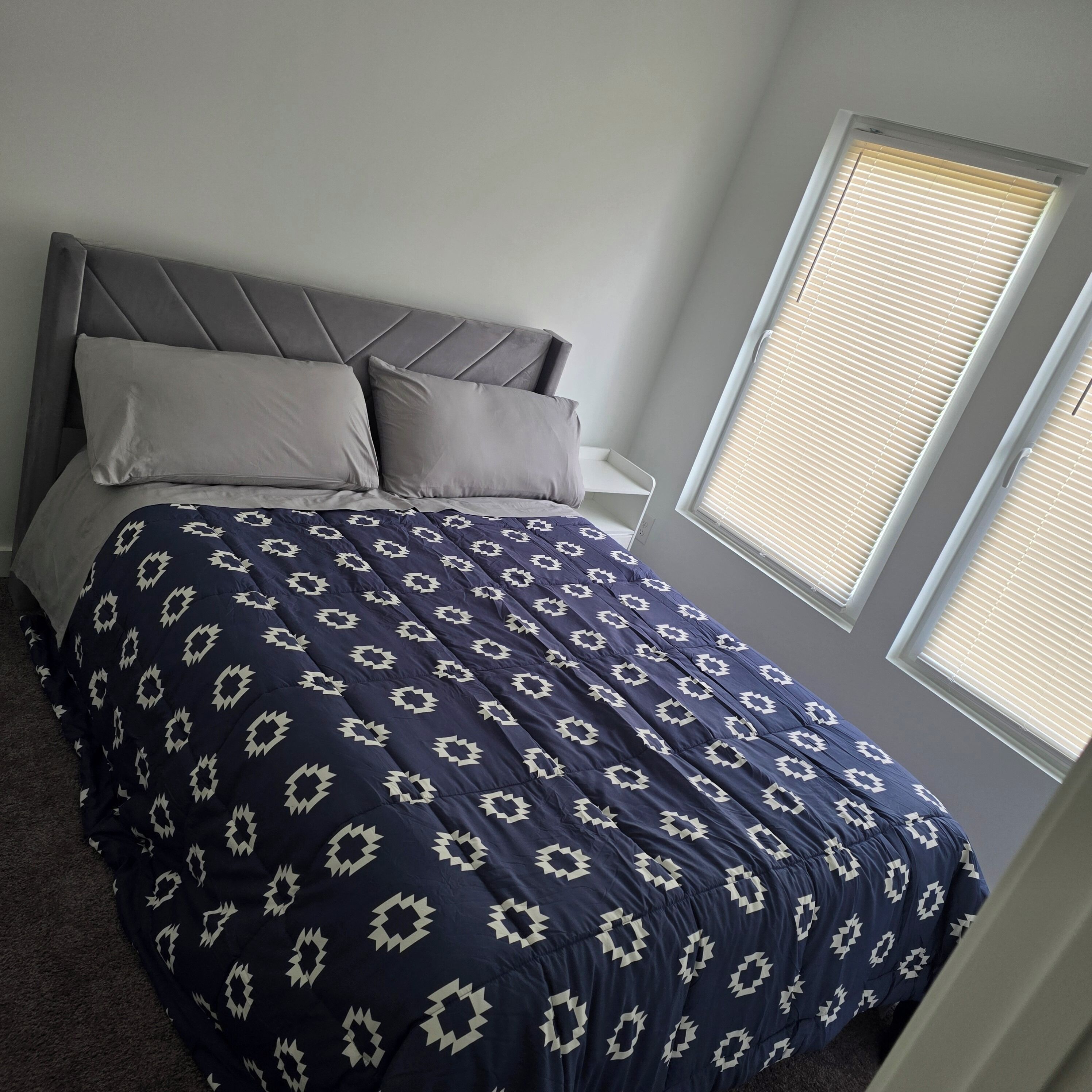 Un dormitorio con cama, cabecera, almohadas y ventana con persianas.