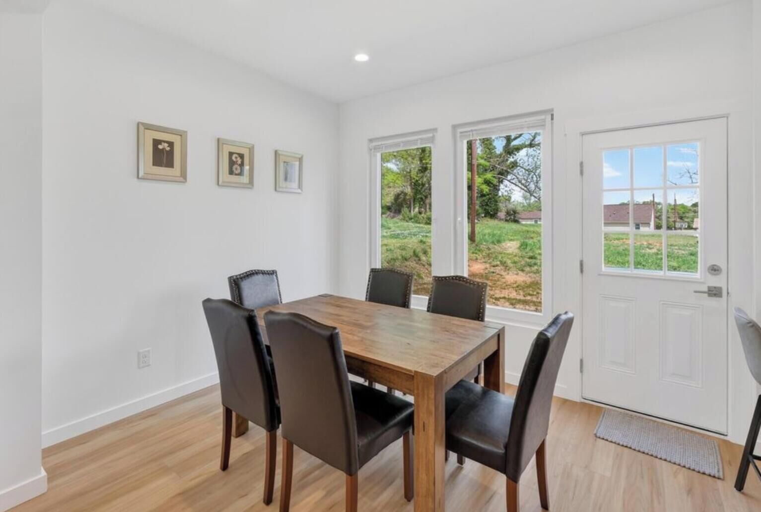 A dining area with a wooden table and six chairs, large windows with a view of greenery, and a white door.