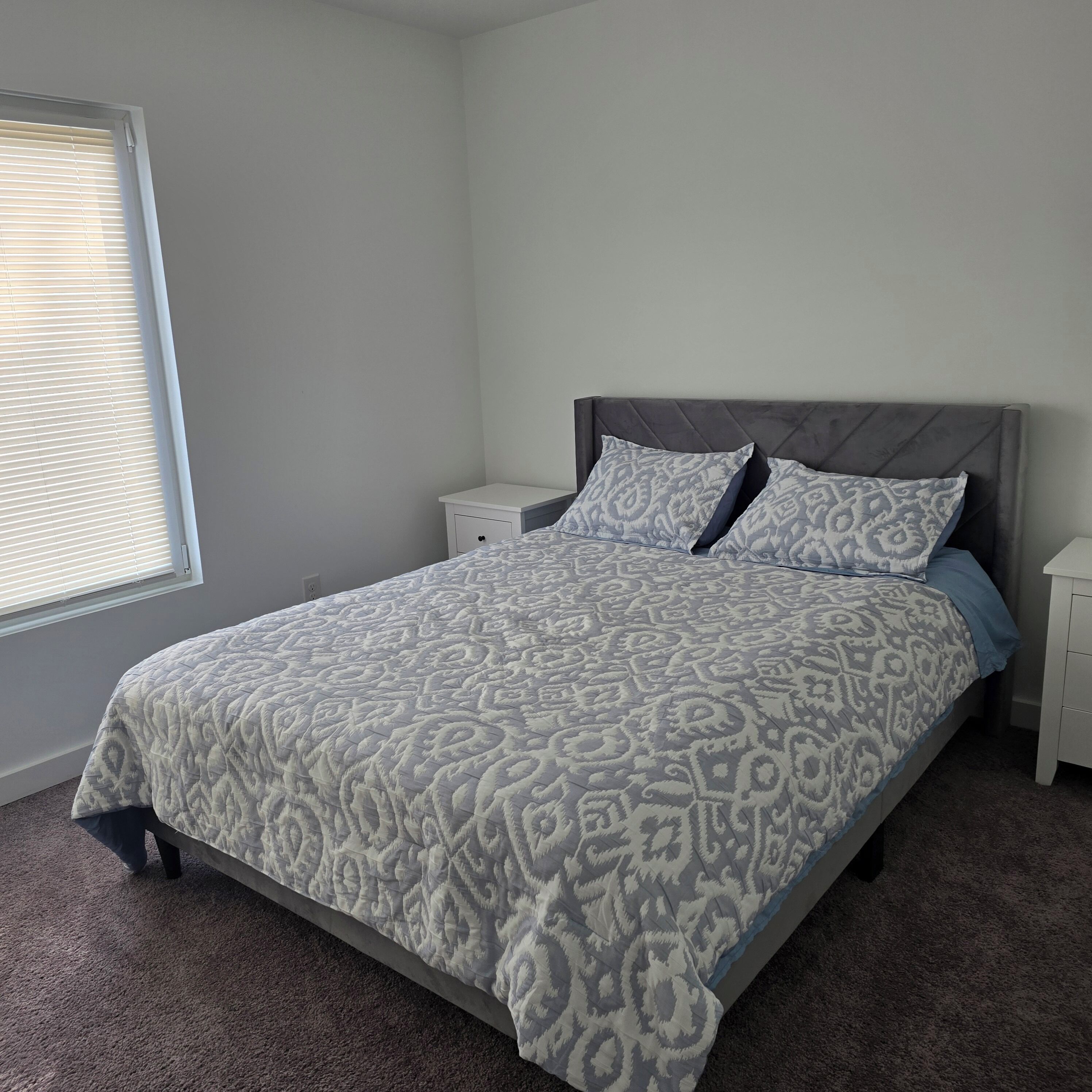 A bedroom with a bed, a nightstand, a window with blinds, and a carpeted floor.