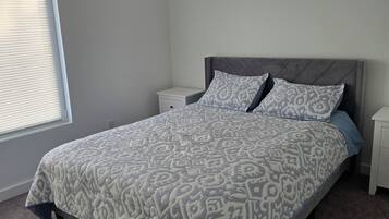 A bedroom with a bed, a nightstand, a window with blinds, and a carpeted floor.