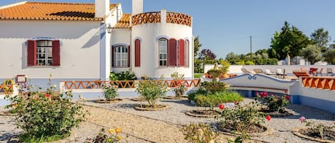 A white building with red shutters and a tiled roof, surrounded by a gravel garden with various plants and flowers.