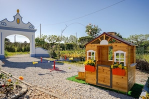 Ein hölzernes Spielhäuschen mit Satteldach und Fenstern auf einem Kiesplatz, mit einem Garten im Hintergrund.