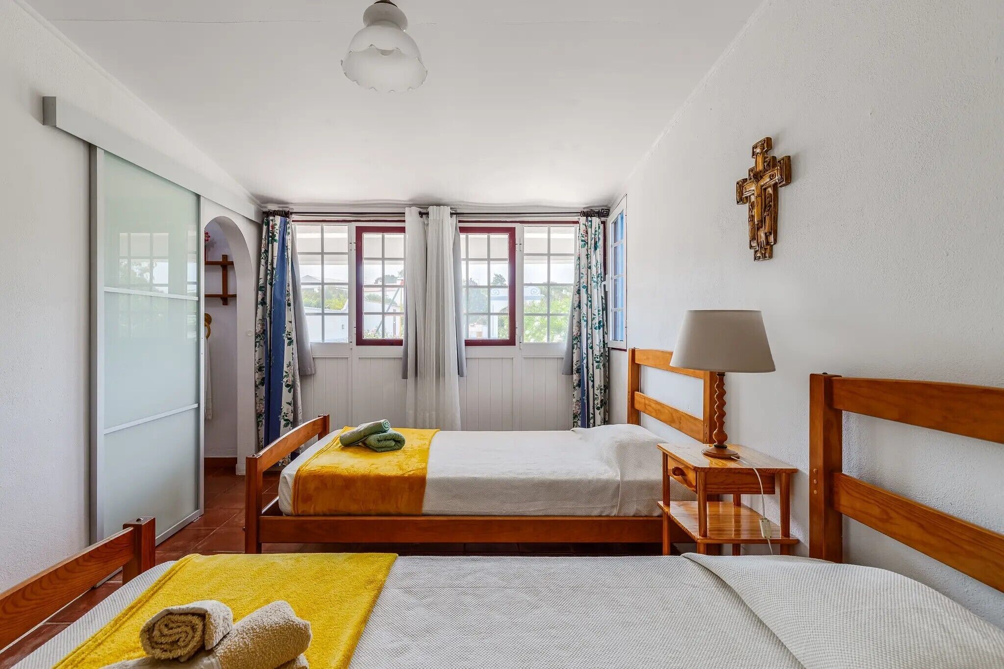 A bedroom with two beds, a wooden headboard, a bedside table with a lamp, a cross on the wall, and a window with curtains.